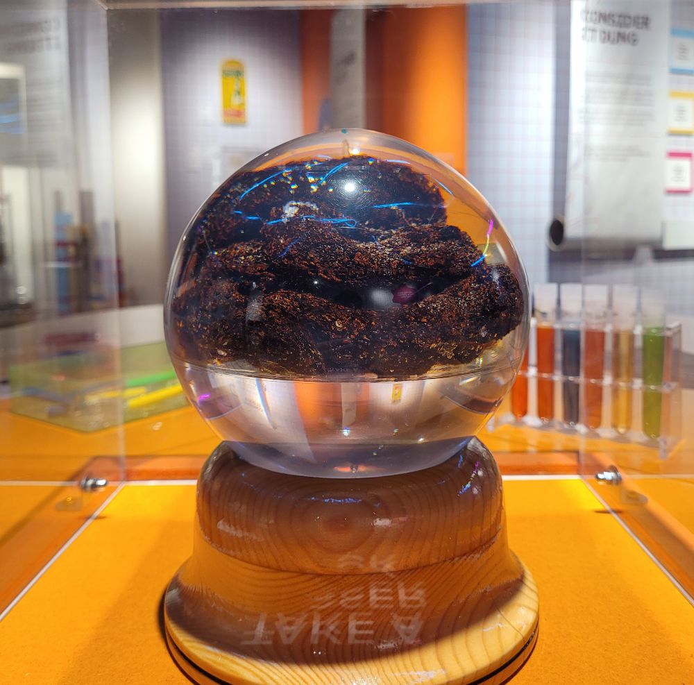 A shiny translucent orb sits on a wooden plinth. Inside is a - frankly enormous - human turd.

In the background there are colourful test tubes and signs, part of a museum exhibition dressed to look like a fun laboratory.