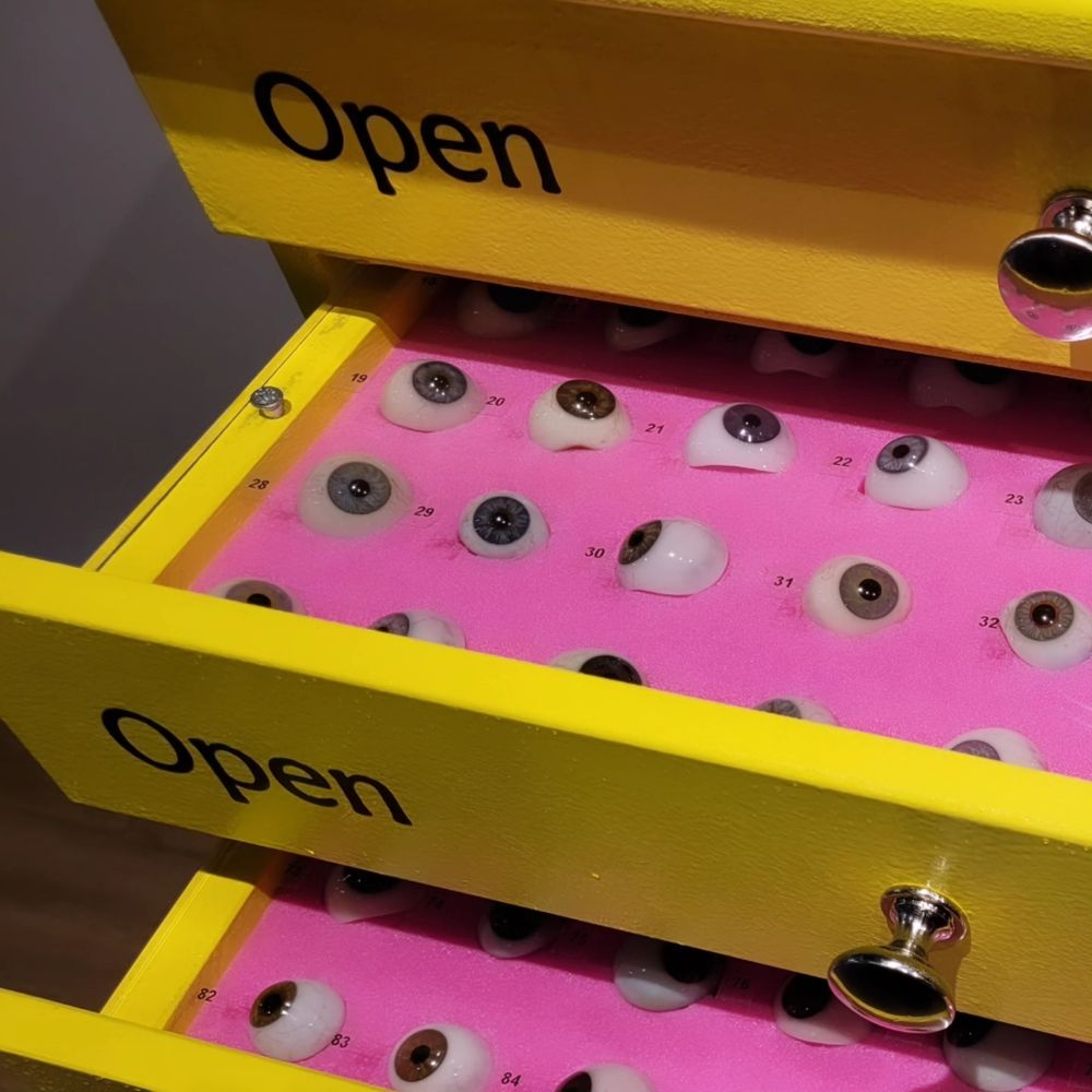 Bright yellow drawers with an inviting "open" label. When pulled open they reveal row upon row of glass eyes sitting on soft pink foam. Each eye has its own unique number to identify it.