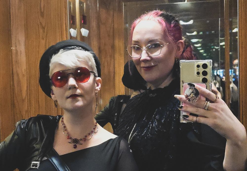 Photo of Marty and I as our vampire larp characters from Darkness Emergent in LA. Marty is blonde with a black beret and red sunglasses. I have pink hair and gold glasses. We're both wearing black.