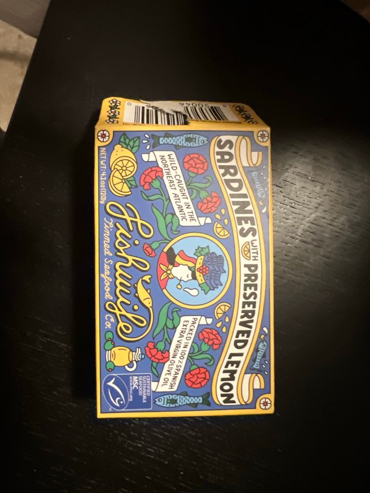 an extremely colorful and decorative box for a tin of fish. Text on box reads “Sardines with preserved lemon. Wild caught in the northeast atlantic. Packed in 100% extra virgin olive oil. FishWife Tinned Seafood Co.” The center of the box holds a cartoon woman smoking a pipe in a red shirt with a yellow ascot, and a hat which is also a basket that holds at least 10 sardines. She looks happy. 