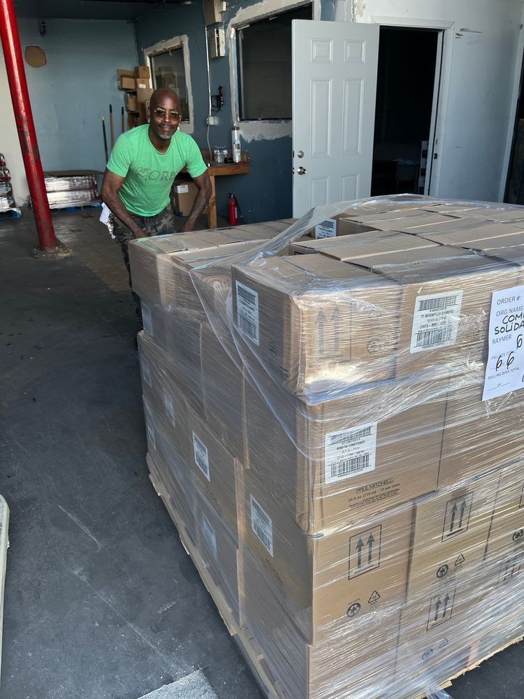 A smiling man in a green T-shirt pushes a pallet of boxes filled with baby supplies.