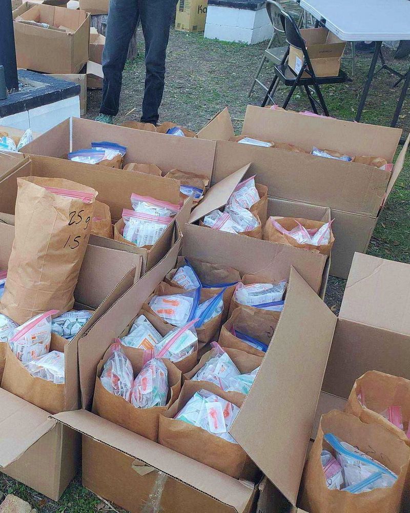 Photo of several cardboard boxes ont he ground outside. The boxes are full of ziplock bags with harm reduction supplies.