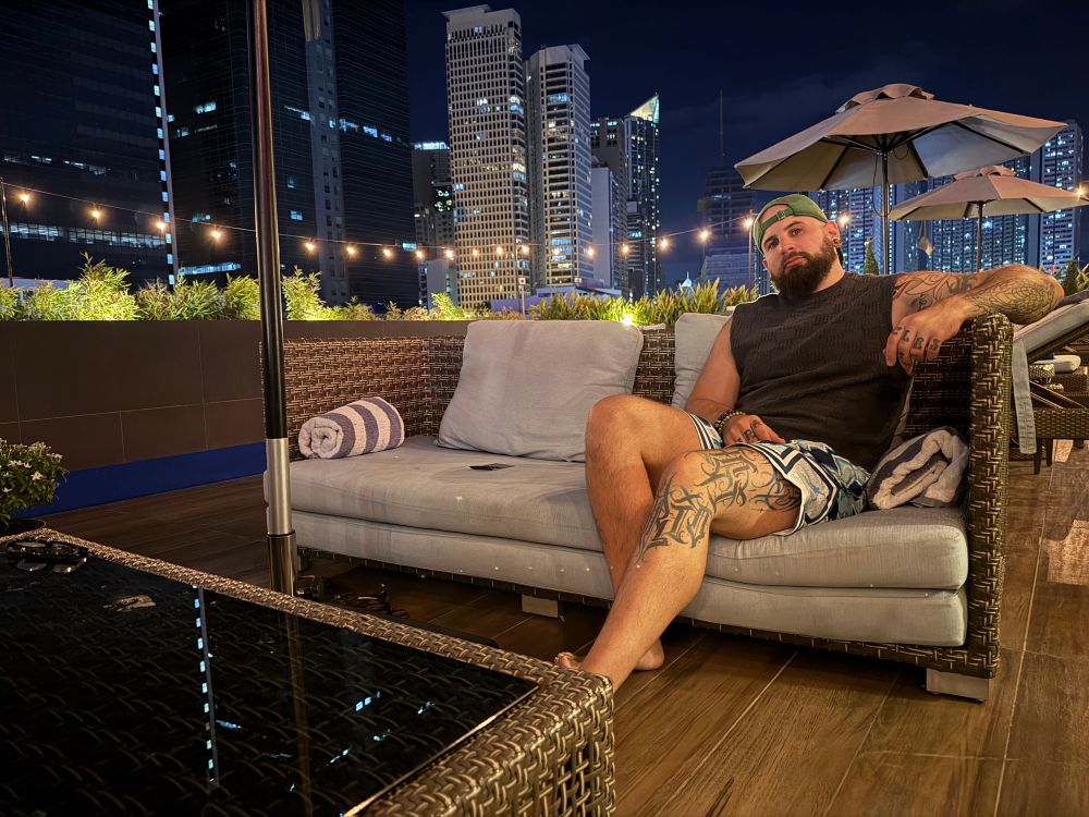 Tattooed bald man lounging on a rooftop at night, showing off tatted thighs and chill Dom energy.