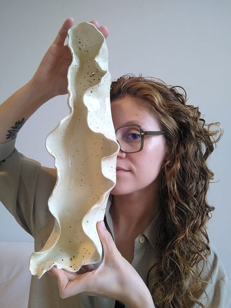Mulher branca de cabelos compridos cacheados em tom castanho claro, usando óculos verde e blusa em tom nude segurando uma peça de cerâmica em tom creme com pintas pequenas pretas e de formato orgânico que remete a forma de corais marítimos.