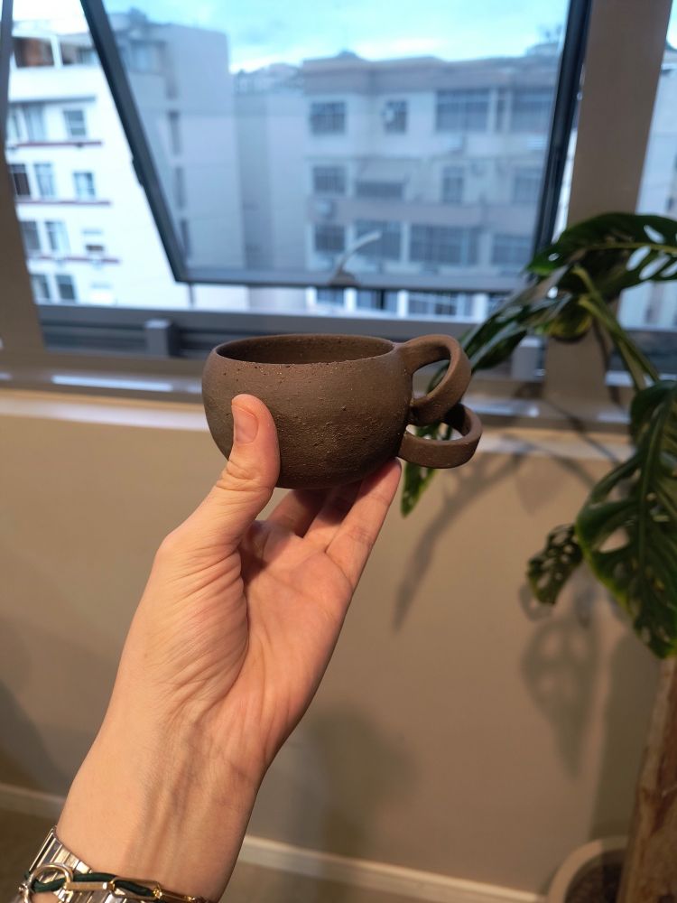 Mão segurando uma xícara feita em argila com alça em formato de um cacho enrolado. Vista frontal.