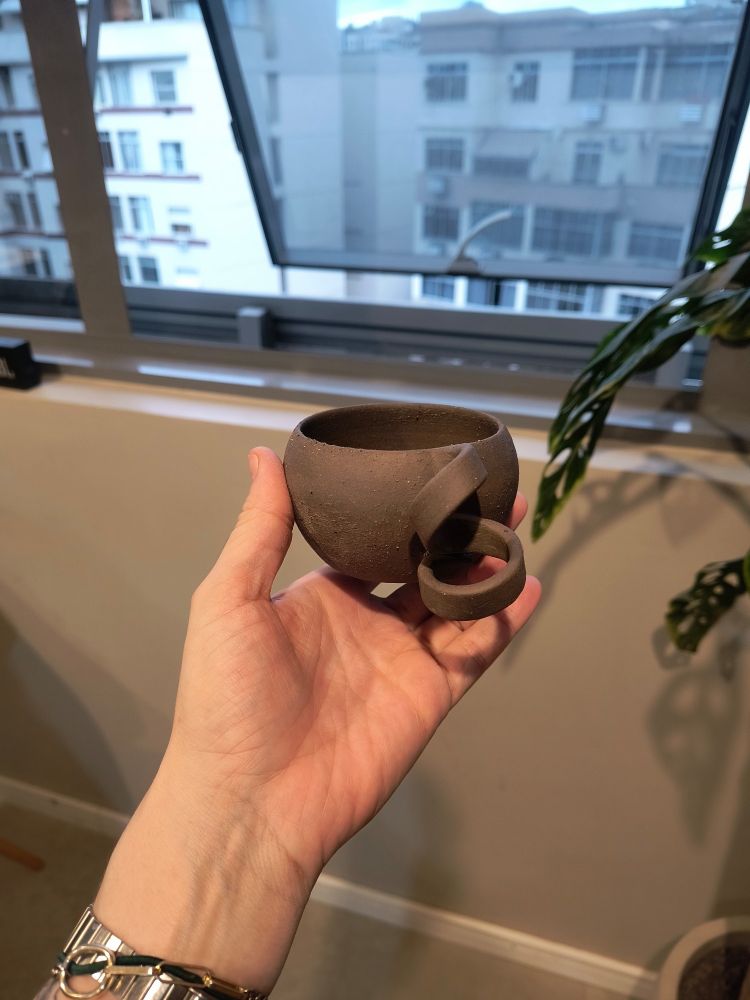 Mão segurando uma xícara feita em argila com alça em formato de um cacho enrolado. Vista lateral com foco na alça.