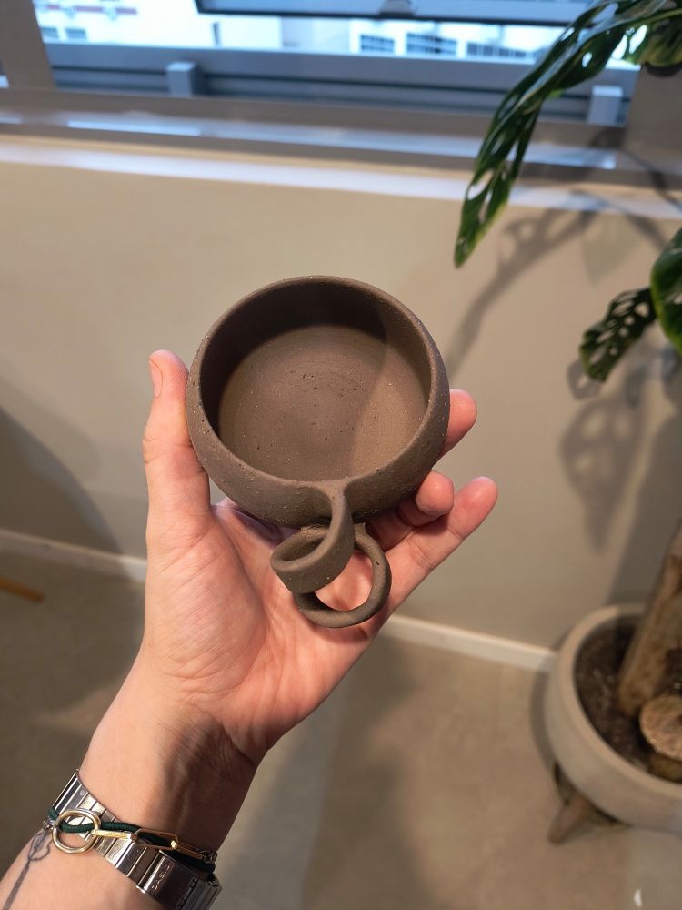 Mão segurando uma xícara feita em argila com alça em formato de um cacho enrolado. Vista superior.