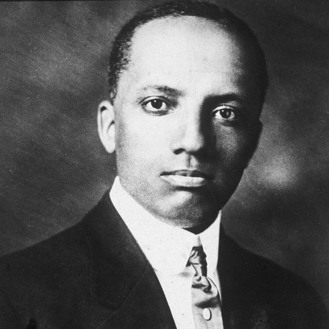 A black and white photo portrait of Carter G. Woodson in a black suit jacket, white collared shirt and tie with an oval tie pin. 
