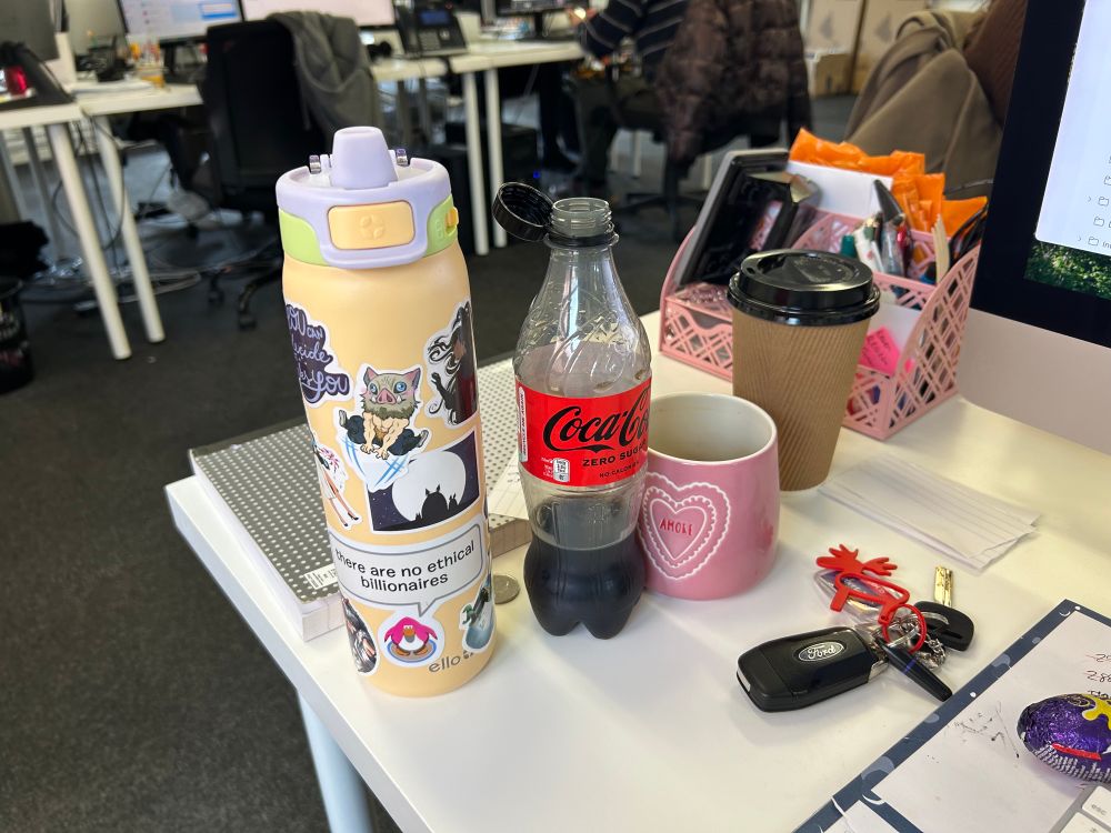a water bottle, coke zero, coffee mug, and disposable coffee cup on a work desk 