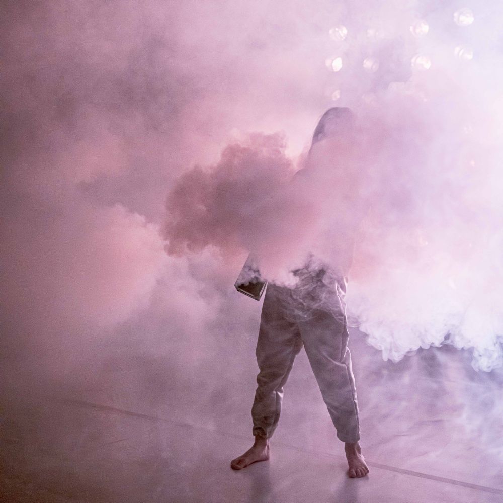 A girl holding a smoke machine wearing a silver jumpuit. The smoke covers her face and body and most of the space arund her. Stage lights are just visible through the smoke behind her.