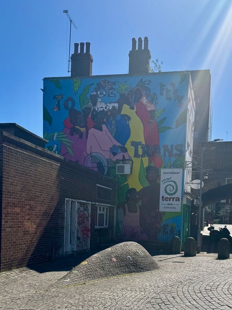Street art in london that reads "to be safely trans," words nestled among a vibrant paintings of black trans people in colorful outfits, on a bright blue background. The art work spans a two story building in a cozy brick alleyway with organic humps of brick sculpted into the walkway with colorful tiles inlaid sporadically.