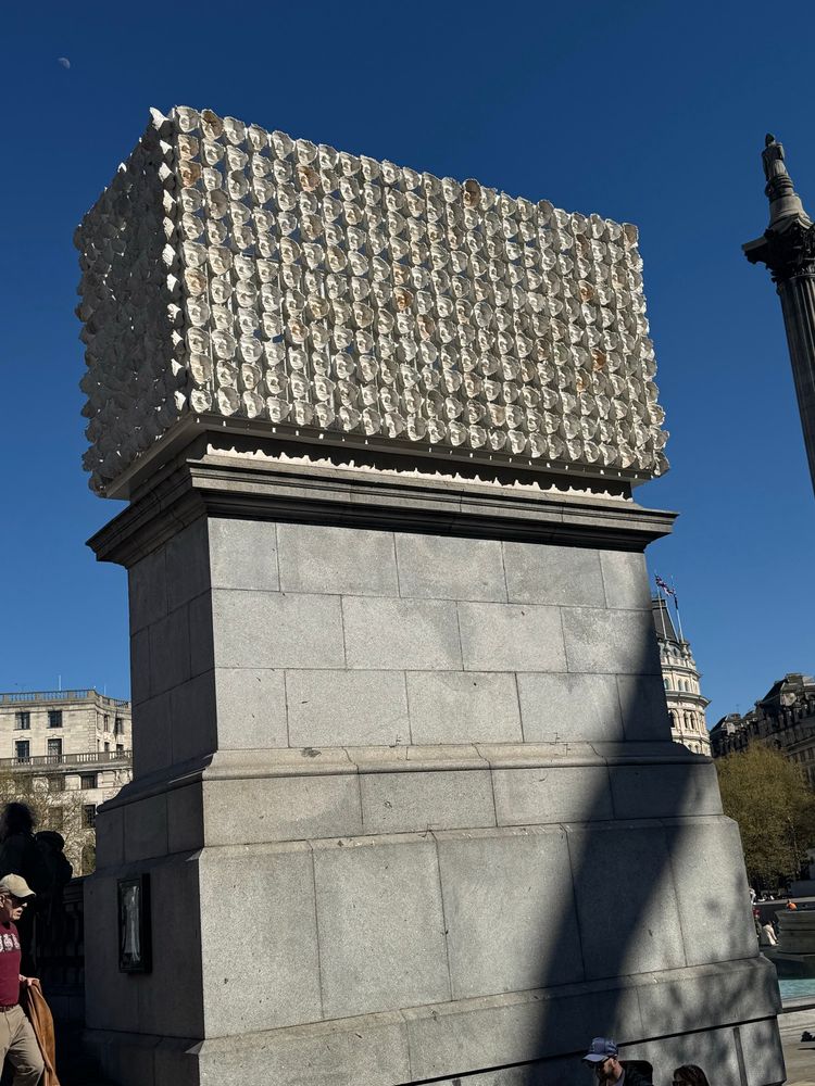 A trans monument in Trafalgar Square made of mouldings of the artist's friend's faces in a rectangular prism, facing inward, on top of a large concrete pylon.