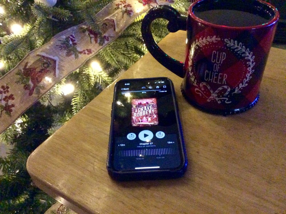 An iPhone sitting on a tv tray top with the audiobook of A Grave Robbery by Deanna Raybourn open on it. Also there is a red and black plaid mug that reads Cup of Cheer in the wreath superimposed over the red and black. There is part of a lit Christmas tree in the background.