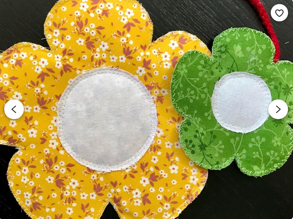 Two flowers made of fabric: The larger one is bright yellow with little brown and white flowers on it and a large white middle (perfect size for a massive mug). The little one is a mossy green, with pale and dark green silhouetted abstract plants. It also has a white middle, and a red cord juts out of it to turn it into a bookmark.