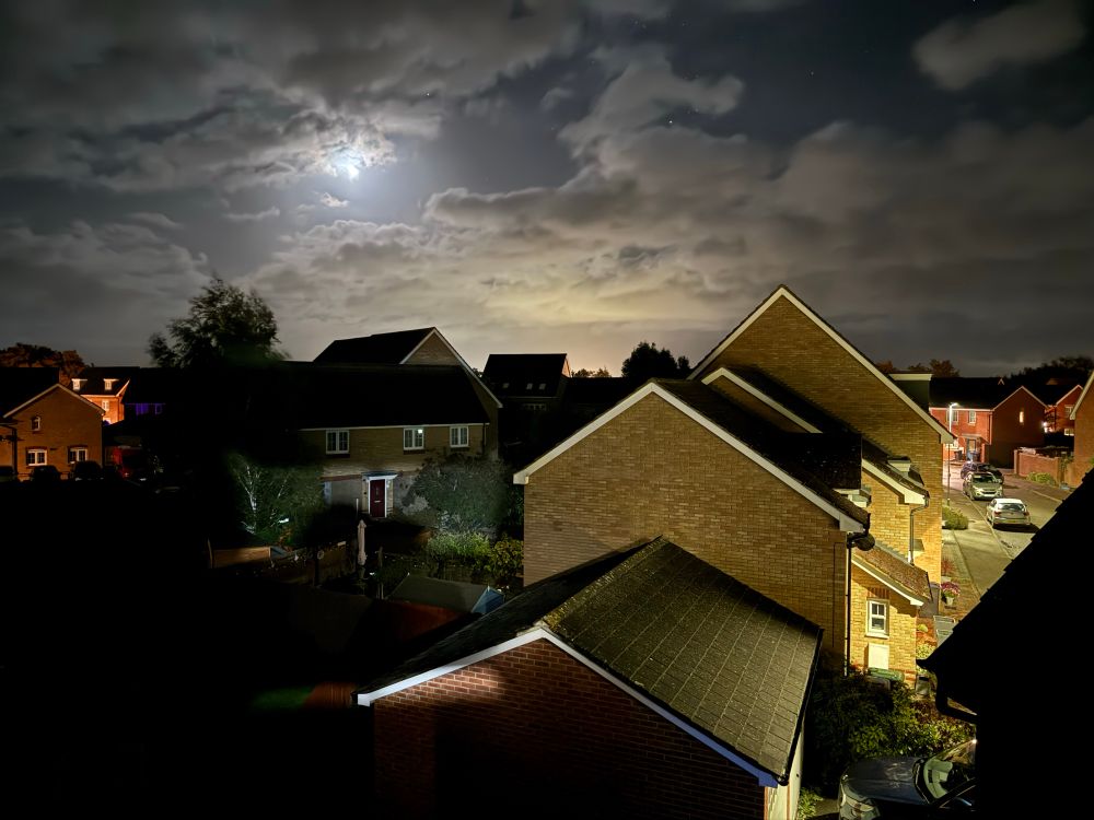 A photo from our top floor window, showing a line of houses along a lit street on the right, a darker area of back gardens on the left and the moon shining through patchy cloud in the sky above.