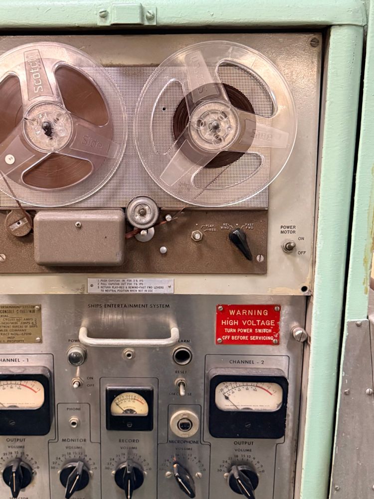 Photo of an old tape deck with multiple dials and switches on it. There are various little readouts and signs, including a bright red one that reads,”Warning High Voltage.” The edges around the tape deck are mint green. 
