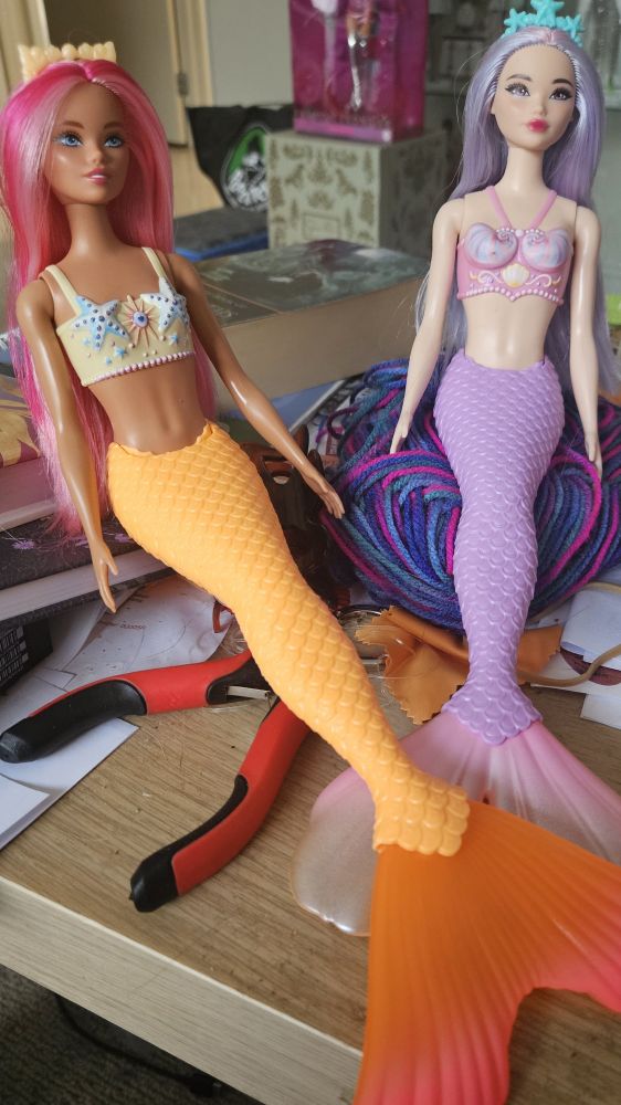 Image is of two Barbie mermaid dolls, one in warm colours, the other in cool colours. Their original headbands have been swapped. They are sitting on clutter on a small beech effect Ikea Malm coffee table  