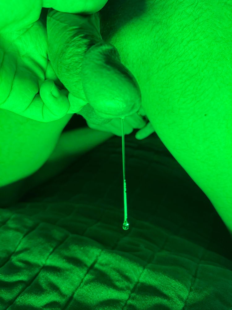 A naked photo of me, bathed in green light. My curved, erect dick is held by a partner's hand, aimed towards the camera. Oozing from the tip is a large drop of precum, dangling inches, nearly touching the sheets below. Behind, my lightly hairy legs.