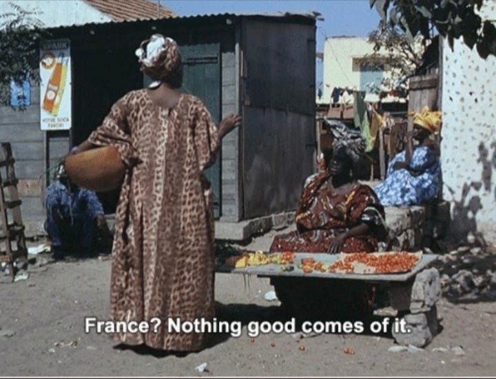 An African woman in a caftan saying “France? Nothing good comes of it.” To a woman selling in a marketplace.