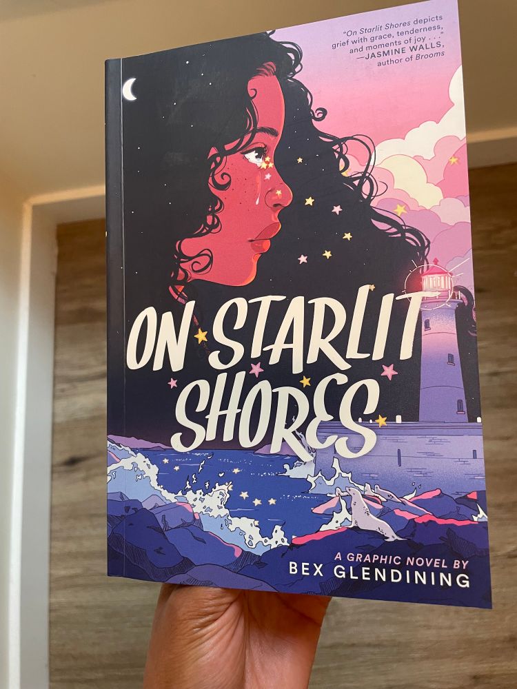 A Black hand holding Bex Glendining's book 'on starlit shores'. The cover depicts a picturesque ocean and sky drawn with pastel colors. A Black girl's face melds with the sky and sea above as she cries literal star tears.