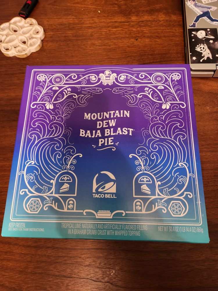 A purple and blue and green blended color box that says "mountain dew Baja blast pie" on it, sitting on a dark brown table