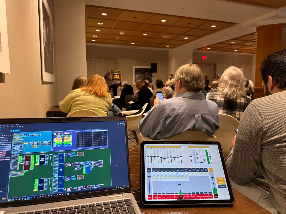 A crowd listens to a reading of The Great Gatsby. In the foreground is an iPad and laptop running the Q-Sys design used for the PA system. 