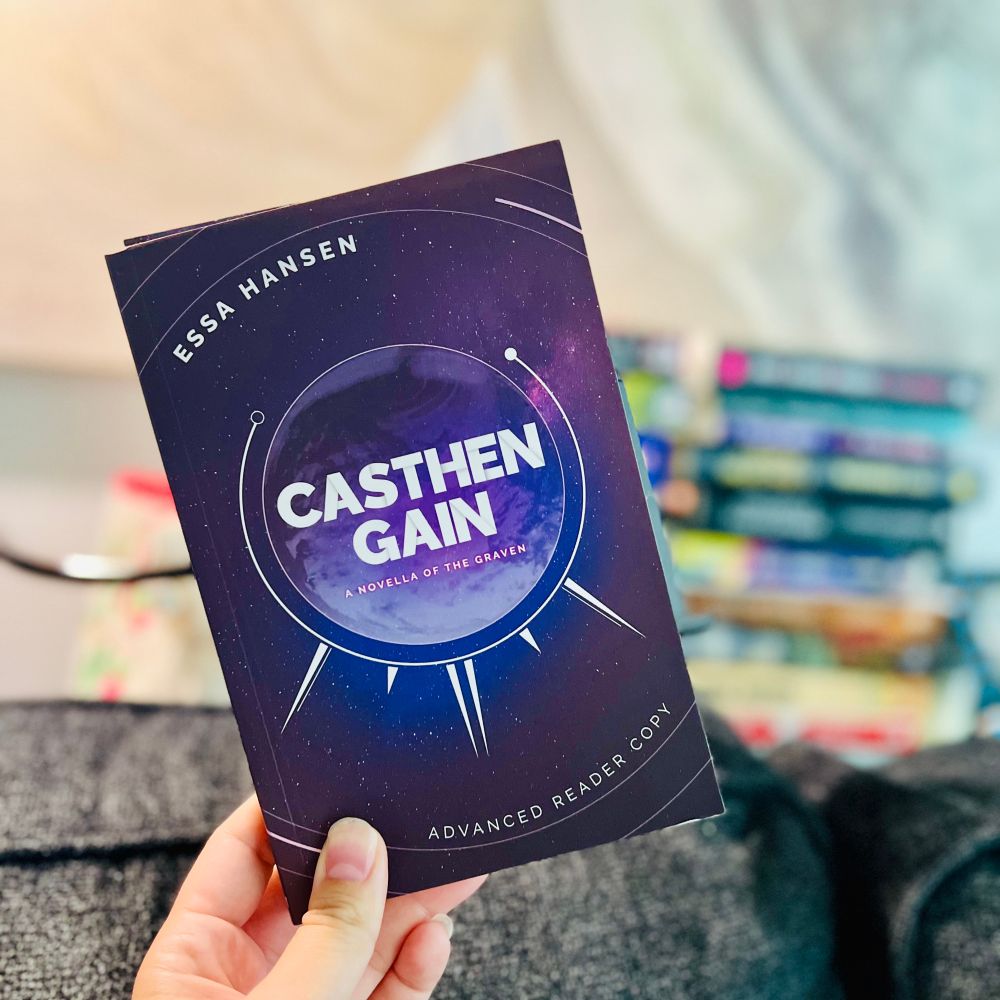 A hand is holding a paperback ARC copy of Casthen Gain by Essa Hansen with a blurry couch and stack of books in the background. 