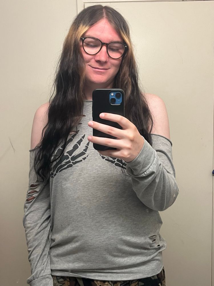 Mirror selfie of tiny visible from the waist up, they’re wearing a grey long sleeve with the shoulders cut off, 