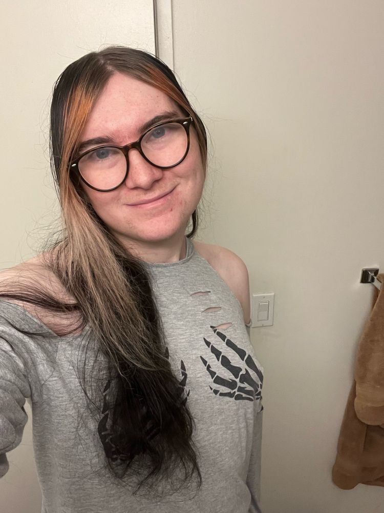 Selfie of tiny, visible from her bellybutton upwards. Holding their phone with her right hand taking the photo. She’s wearing a grey shirt with skull hands over her tits. Her shoulders are exposed, with their hair coming down the right side of her head. She’s smiling while tilting her head slightly 