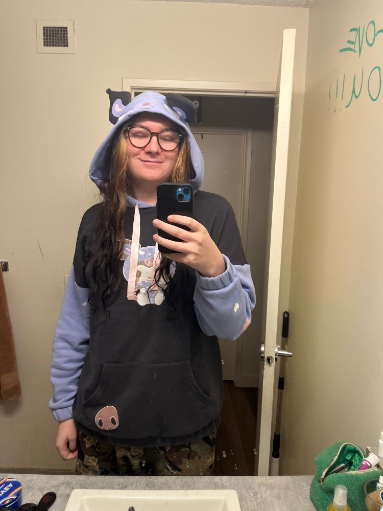 Bathroom mirror selfie of tiny. Visible from the waist up. She’s wearing a oversized hoodie with the hood up, her long hair flows down each side of her face smiling at you 