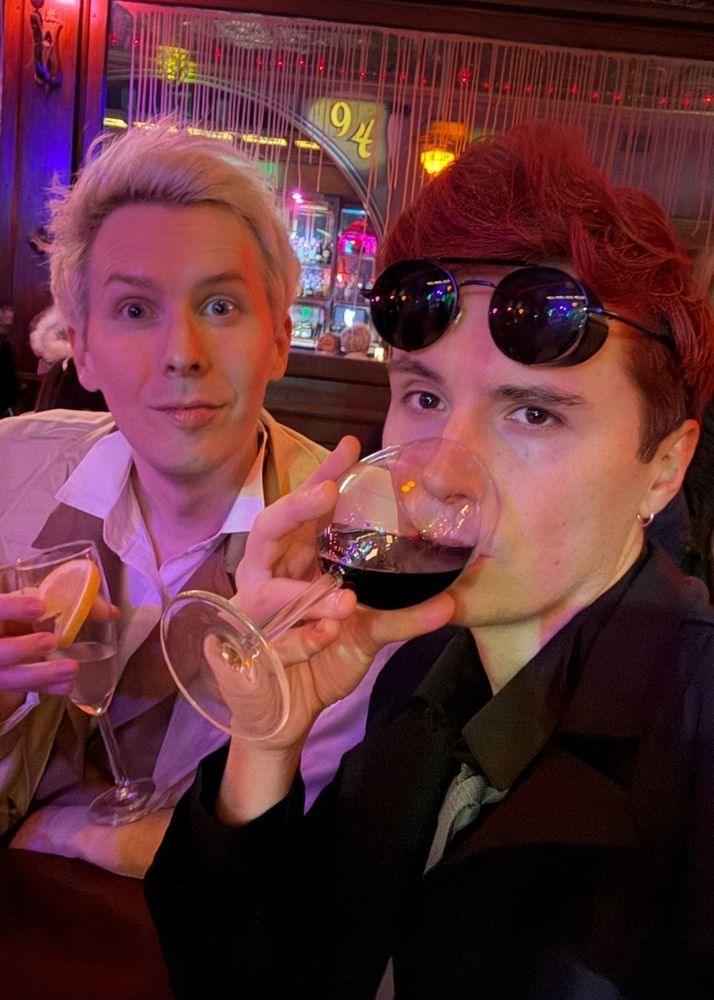 dan and phil selfie from Halloween 2024. phil is dressed as aziraphale and dan as crowley. they're both holding a drink in hand. dan is taking a sip of his drink.