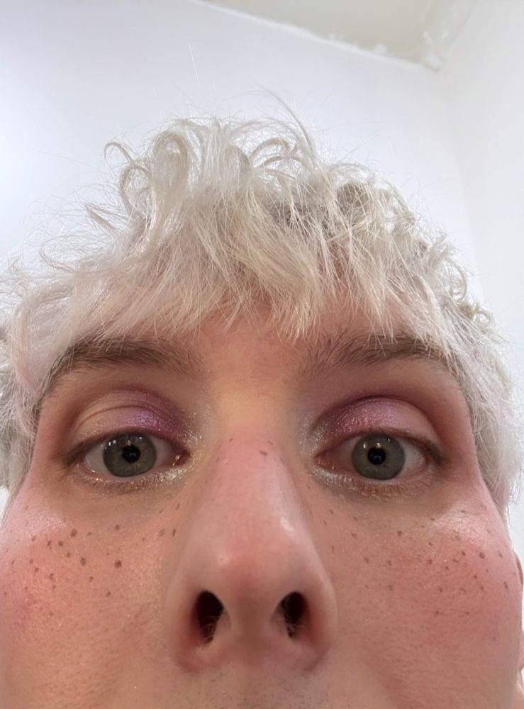 selfie of phil where only the top half of his face is visible. he's wearing some pink blush and has some drawn on freckles on his cheeks. he's wearing a pink eyeshadow with some white in the inner corners of his eyes.