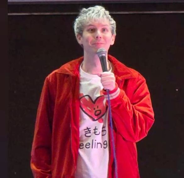 phil lester holding a microphone. he's wearing a white shirt with a red heart on it and some text underneath, and a red jacket on top. he's smiling.