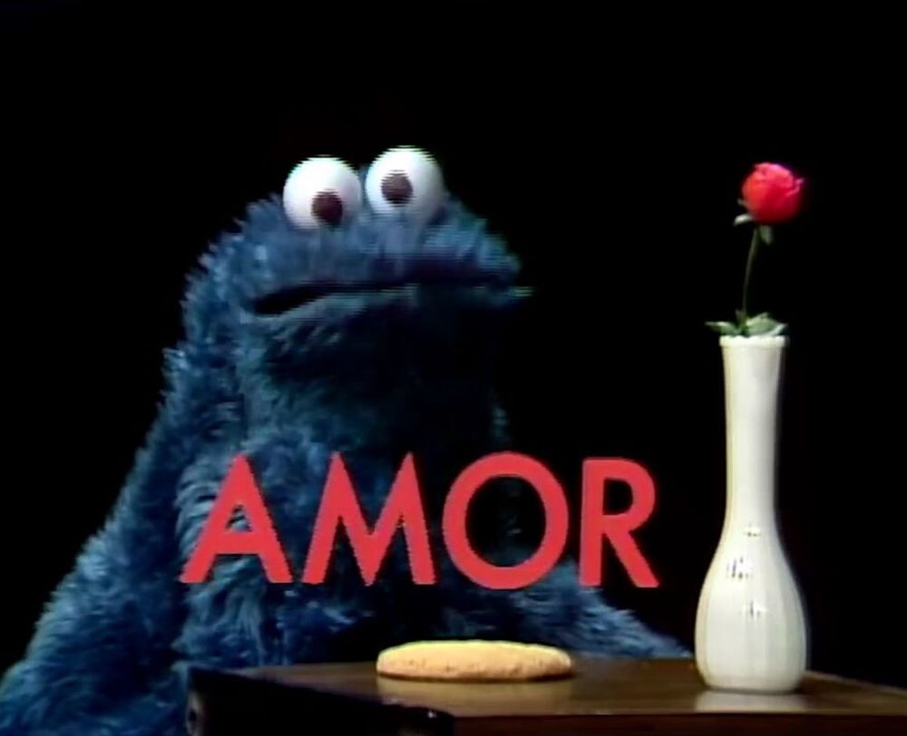 Cookie Monster sitting at a wooden table on a black background. On the table rests a cookie and a slim white vase holding a single rose. One word, AMOR, in red across the scene. 