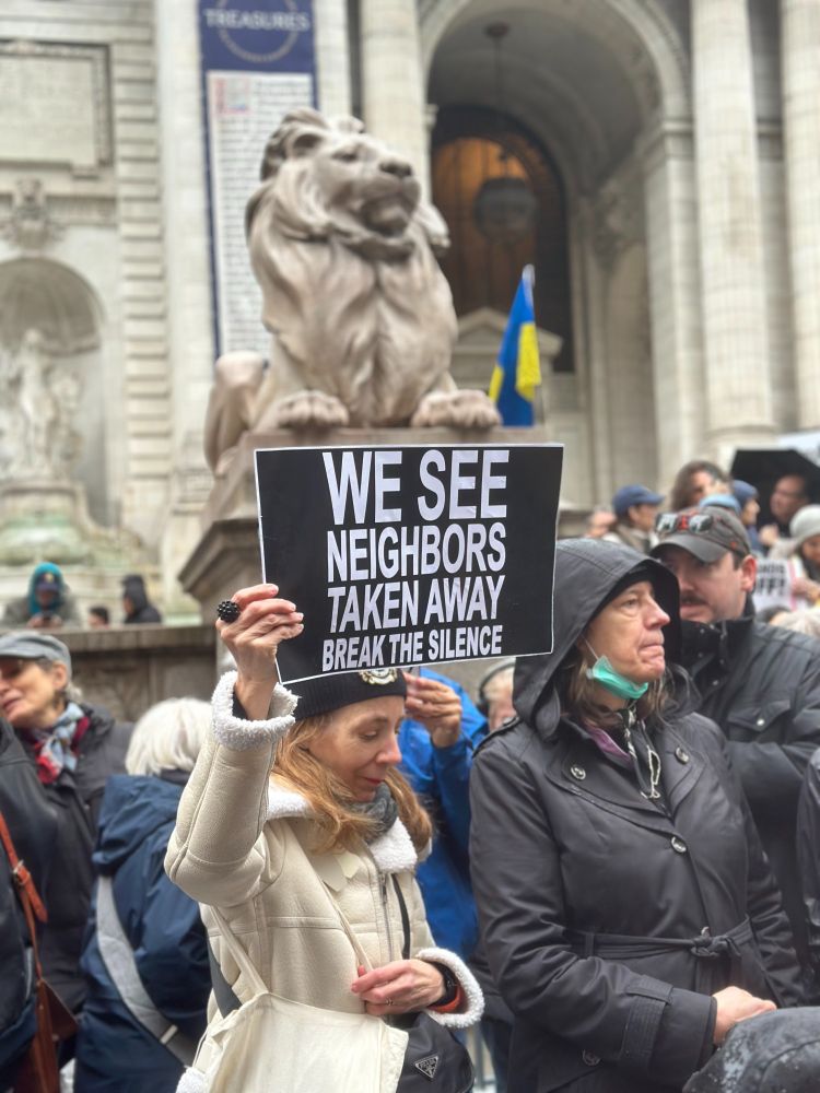 A woman at the Hands Off march of April 5, 2025, holding a sign saying "WE SEE NEIGHBORS TAKEN AWAY BREAK THE SILENCE" in front of one of the stone lions in front of the New York Public Library. A Ukrainian flag flies in the background.