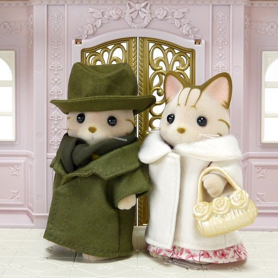 Two cat calico critters in front of a dollhouse with a fancy golden gate. Both critters are lightly cream with brown stripes. On the left is a critter dressed in a green hat, matching green overcoat, and a darker green scarf. The critter is posed to look as though it is holding hands with the critter on the right, which is dressed in a long white coat with fur trim and a pink floral dress, and it is holding a plastic handbag.