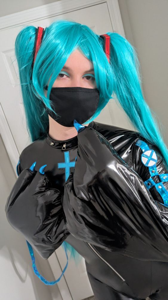 Natalie, dressed again in the Hatsune Miku jumpsuit, with wig, facemask, and make-up. The shot is relatively close-up, Natalie staring right into the camera. One of her paws is on her chest, with the other curled with a claw on her chin, like thinking or teasing.