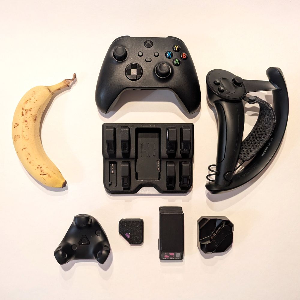 Showcase of various objects on a white background from a top-down view. Objects are a FluxPose dock populated with 7 trackers, a separate FluxPose tracker, a FluxPose beacon, a Tundra tracker, A Vive Tracker 3.0, an XBOX Series controller, a Valve Knuckle controller and a banana. The FluxPose tracker is significantly smaller than all other objects. The beacon is slimmer but longer than the Tundra tracker and about the same length as the longest side of the Vive Tracker 3.0. The dock has about the same footprint as the XBOX Series controller.