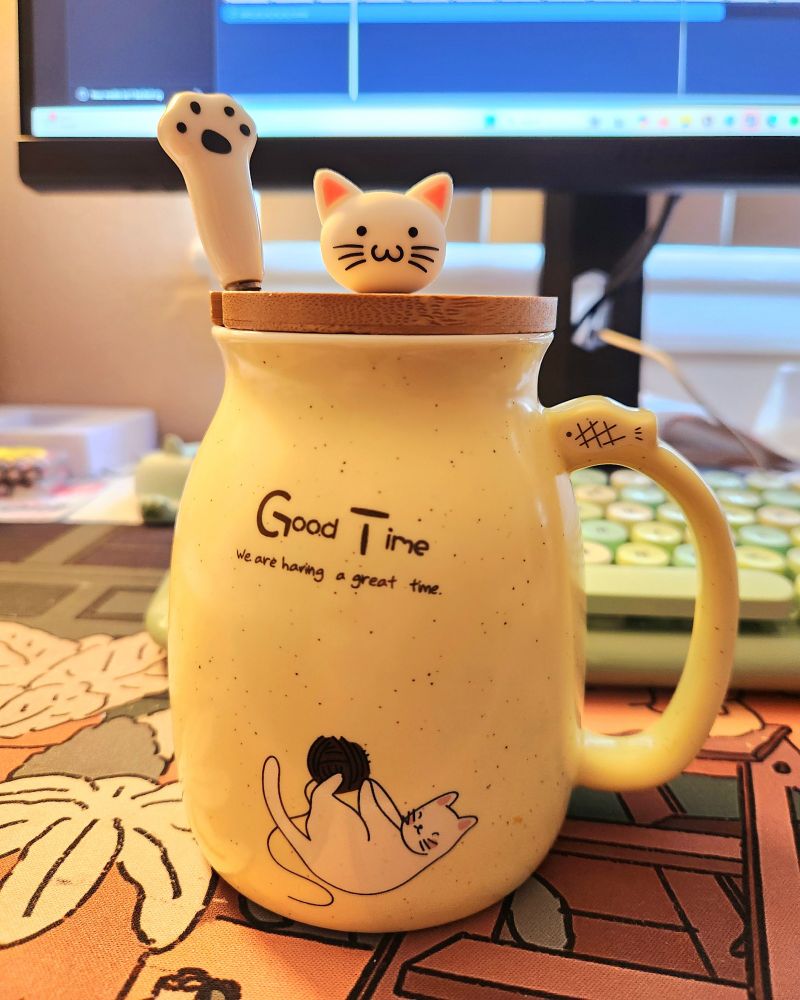 Yellow mug sat in front of a keyboard and computer monitor. On the mug is an illustration of a cat playing with string. There is text on the mug that says "Good Time. We are having a great time." There is a wooden lid on top that keeps the coffee warm with a knob on top in the shape of a white cat head. There is a hole cut into the lid to make room for a tea spoon. Sticking out of the mug is a tea spoon designed in the shape of a white cat paw.