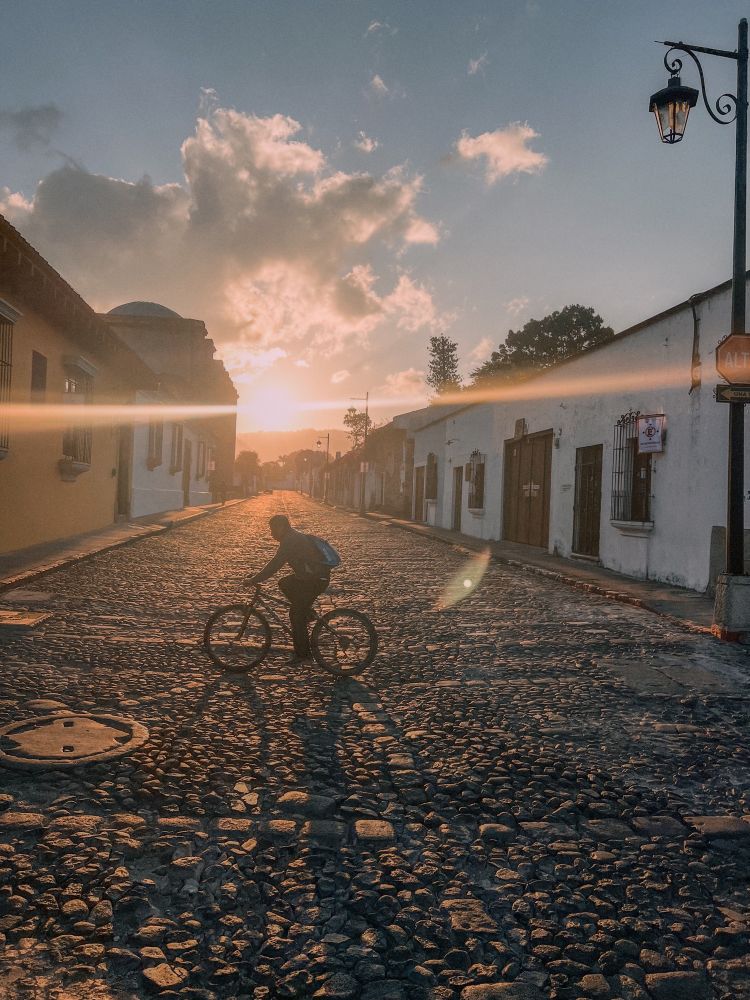 A cyclist rides across a cobblestone street at sunset, framed by long golden rays of sunlight. Colonial houses line both sides of the street, their white and yellow walls glowing in the fading light. A warm, cinematic atmosphere fills the scene.