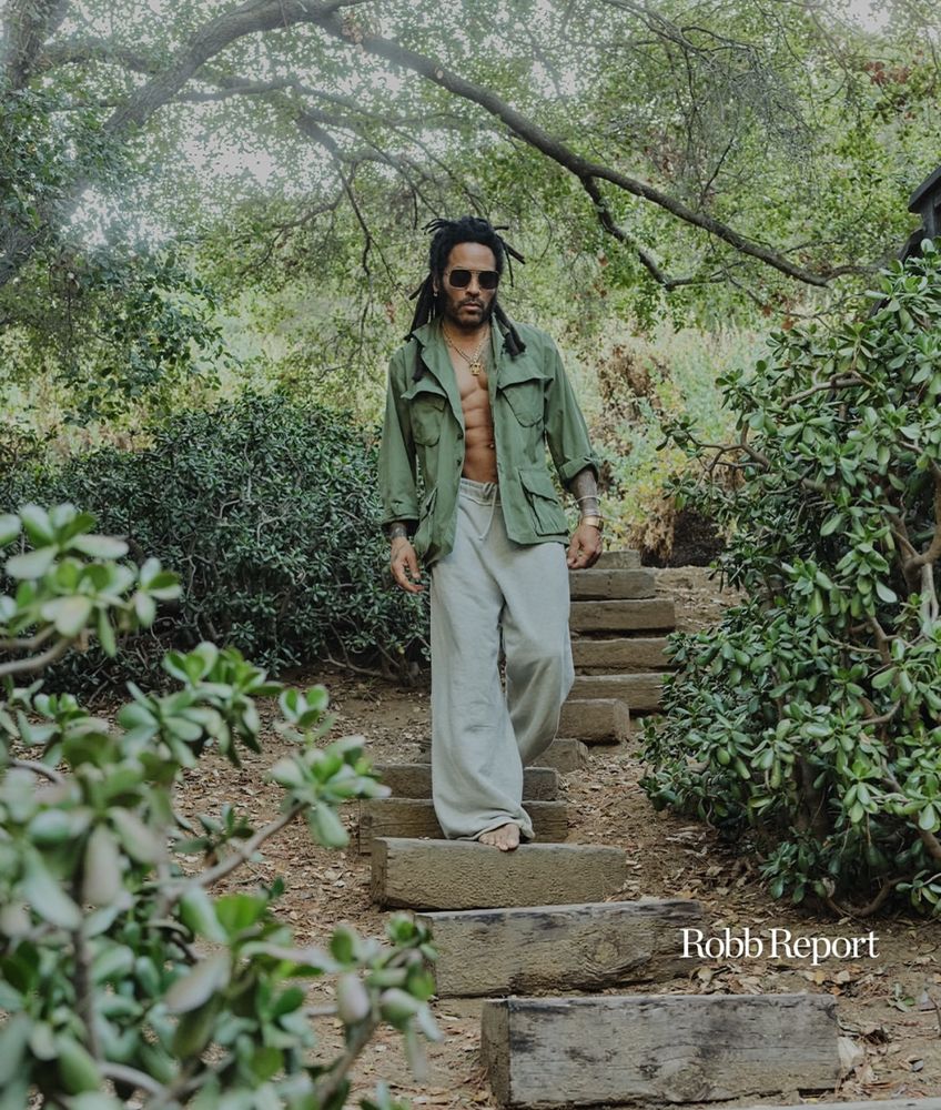 Lenny Kravitz walks barefoot down rustic wooden steps in a lush, shaded garden. He wears loose gray pants and an open olive-green military jacket revealing layered necklaces and a bare chest. Sunlight filters through the trees above, creating a tranquil, earthy atmosphere.
