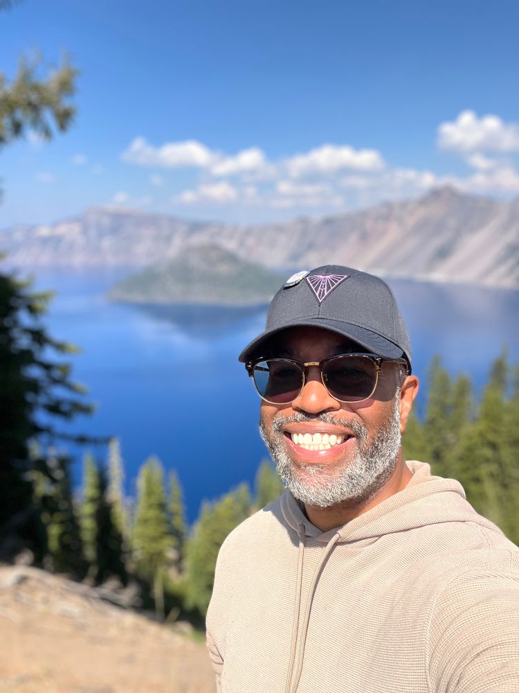 A man smiles brightly in a selfie, wearing sunglasses, a gray cap, and a beige hoodie. Behind him, Crater Lake’s deep blue water glistens beneath a clear sky, framed by evergreen trees and rugged cliffs.