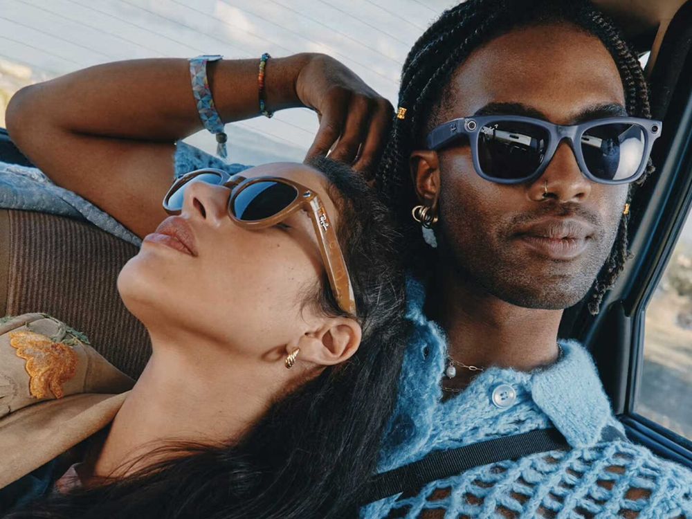 Two people sitting together in the backseat of a car, both wearing Ray-Ban smart sunglasses with built-in cameras. The woman leans back resting on the man’s shoulder, wearing light brown frames, while the man looks forward with navy frames, braided hair, and gold jewelry, dressed in a light blue knit sweater.