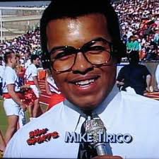 A young male sportscaster wearing large glasses, a white shirt, and tie holds a microphone labeled “Sun Sports.” The text on screen reads “Mike Tirico.” Behind him, cheerleaders and a stadium crowd are visible on a sunny day.