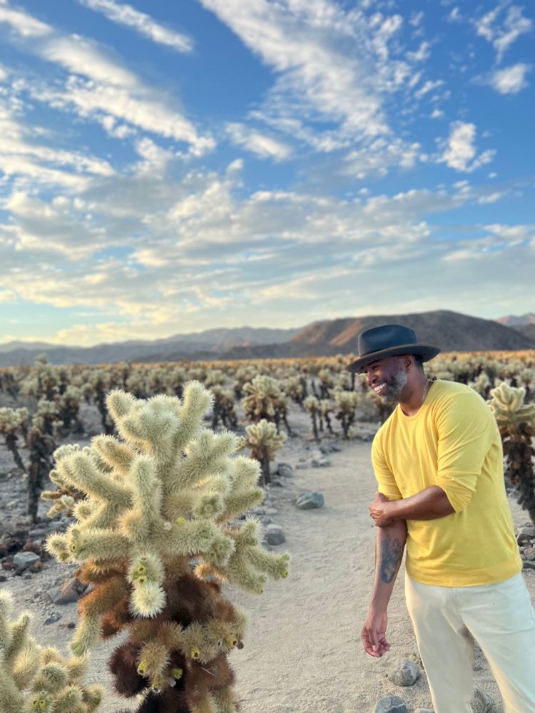 A man in a mustard yellow shirt and cream pants stands smiling among cholla cacti under a bright morning sky scattered with fluffy white clouds. The desert mountains form a hazy backdrop.