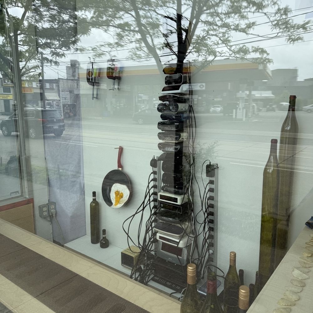 A storefront window display features a stack of electronic devices surrounded by kitchen items and empty wine bottles.