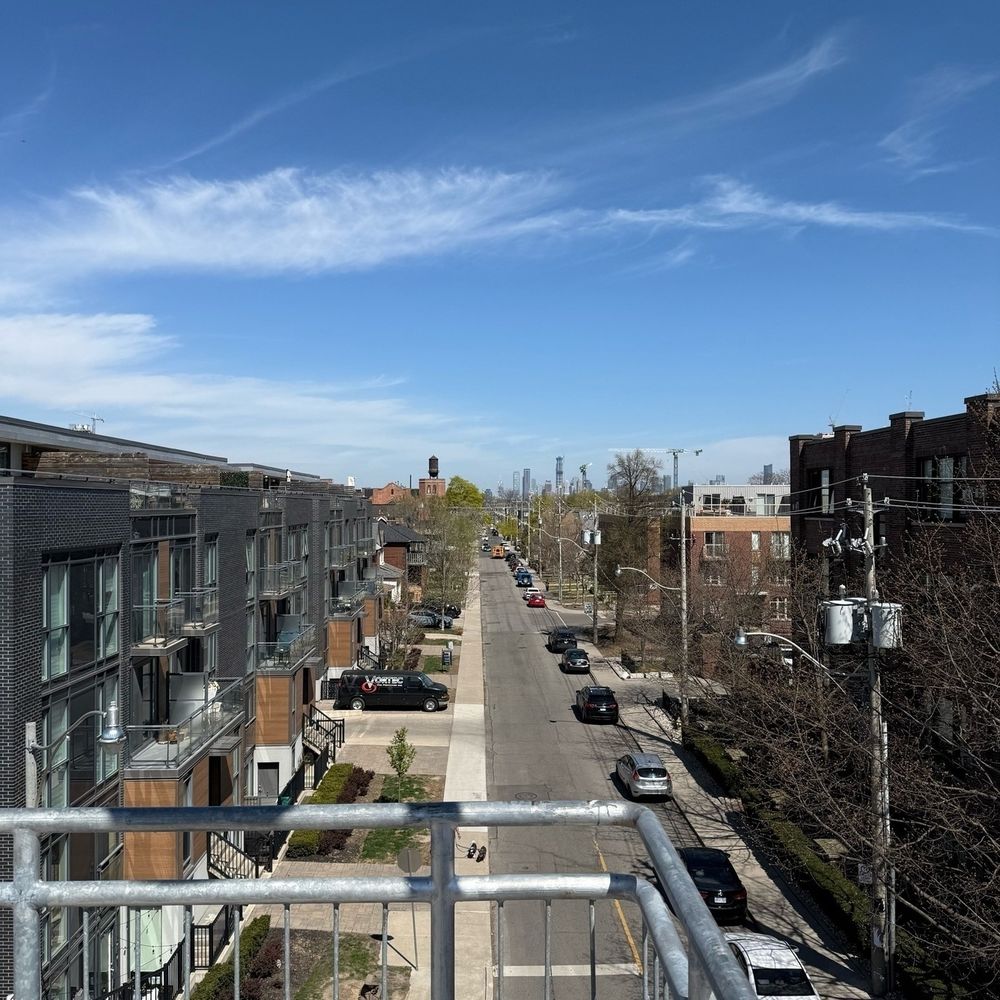 This photo shows a sunny spring day in a quiet Toronto neighborhood, with a straight street lined by low-rise buildings and parked cars. Trees are just starting to bud, and the distant skyline, including the CN Tower, is visible under a bright blue sky.