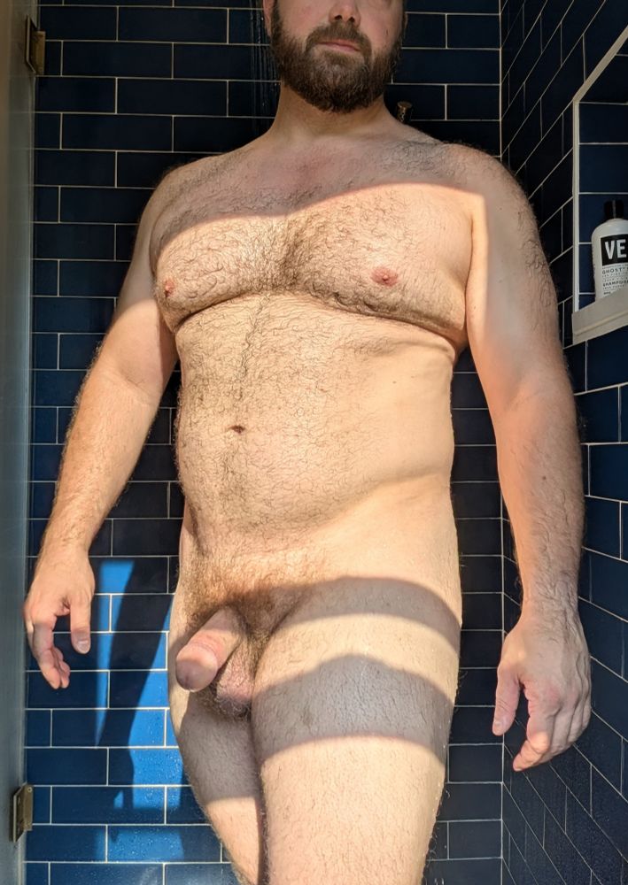 A hairy man stands nude in a blue tiled shower. He is nude. The water is running behind him.