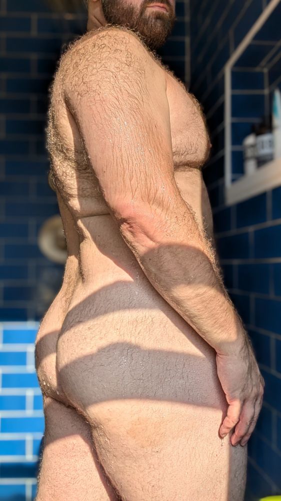 A man stands in side profile in a blue shower. The camera is focused on the man, so the shower is blurred. The man's hairy body and thick legs, however, are in sharp focus.