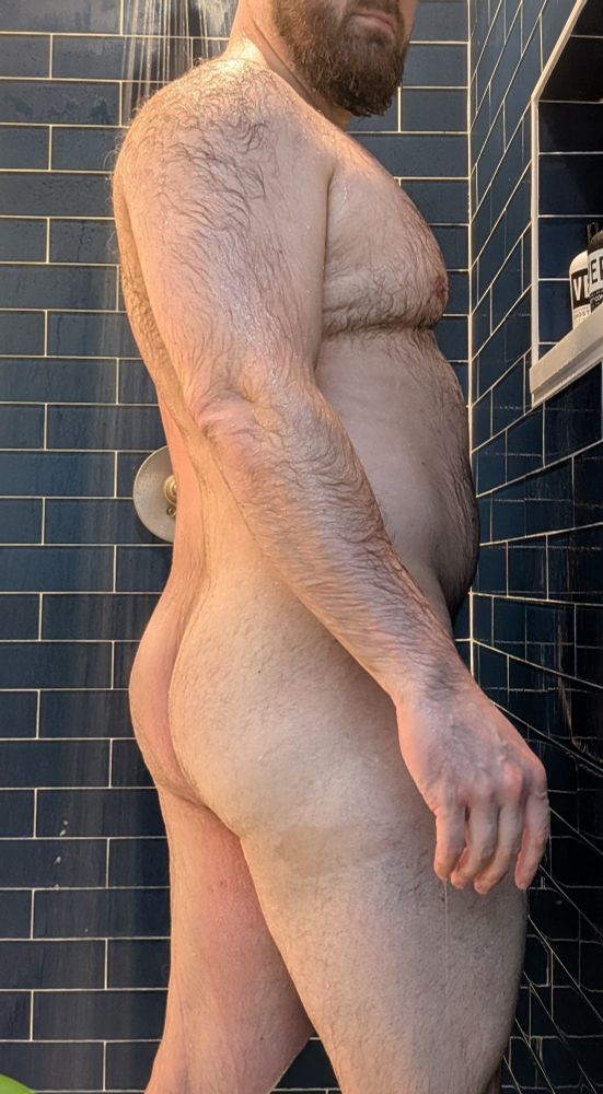 A man stands in a blue shower in profile. You can see his butt. He is wet.